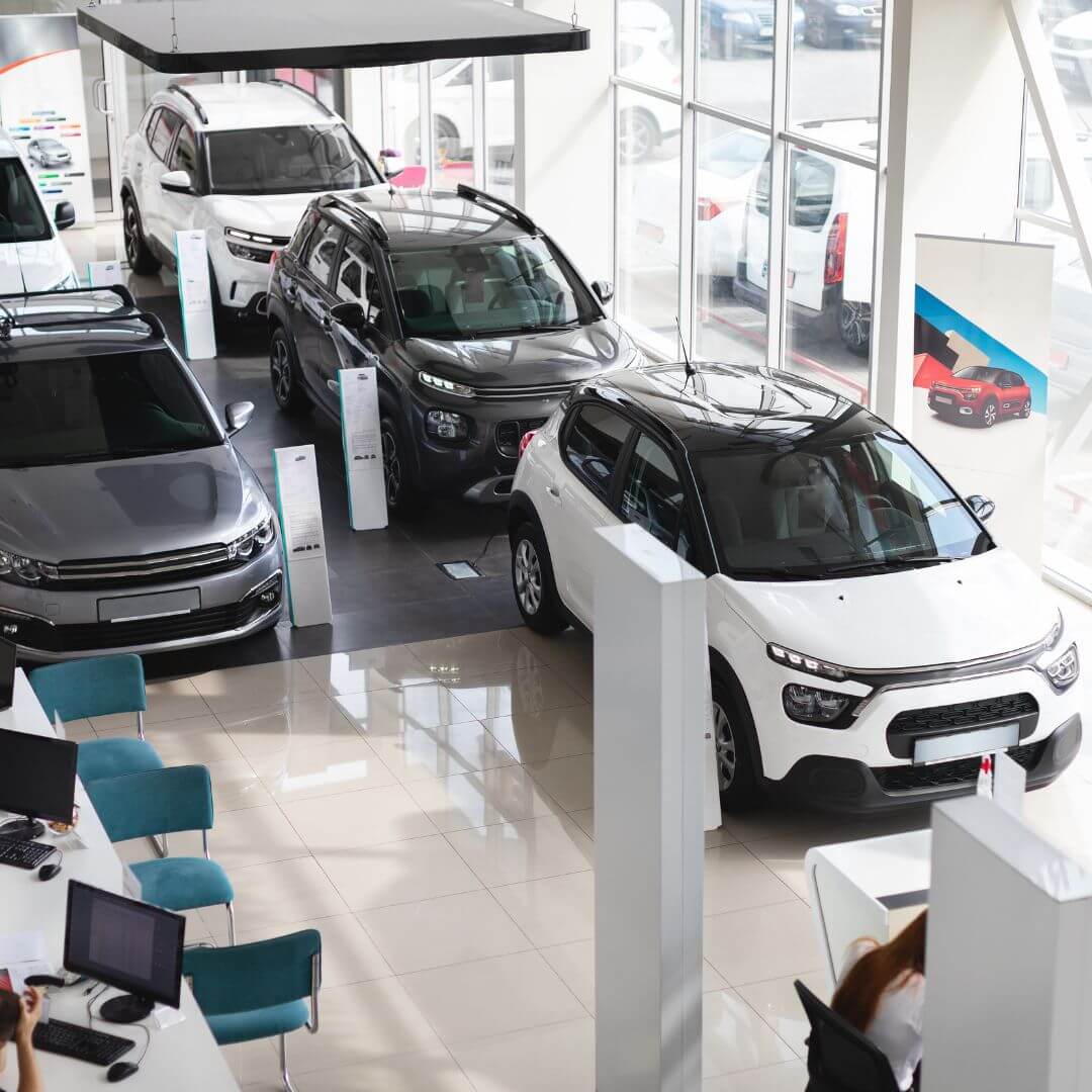 High-angle view of a car dealership showroom with polished white tile floors, featuring various clean cars and blue chairs at consultation desks.