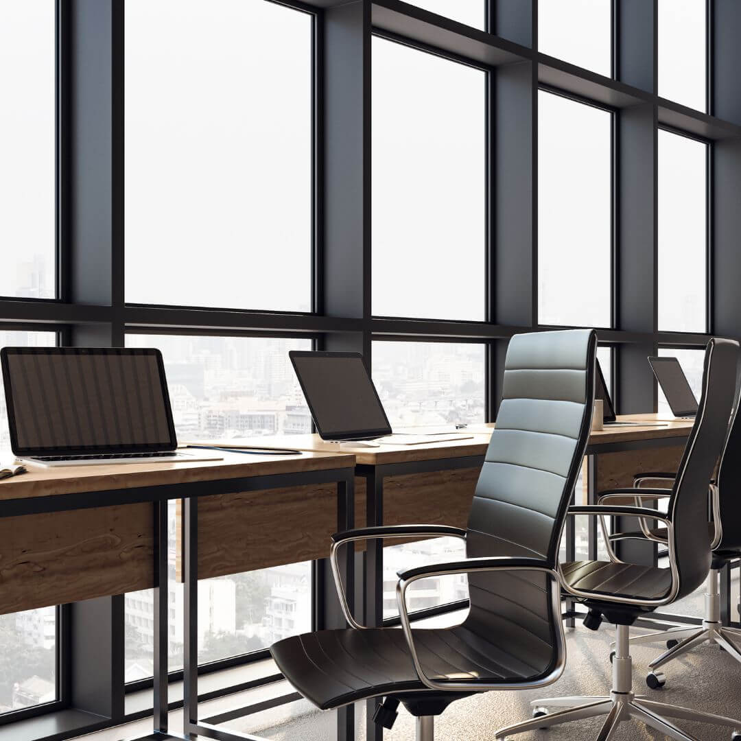 Close-up of a clean office workstation featuring wooden desks, black ergonomic chairs, and laptops in front of large windows with a city view.